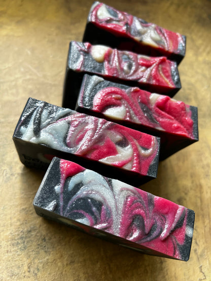 Five rectangular handmade soap bars with a marbled swirl pattern of red, pink, white, and black, arranged diagonally on a textured brown surface.