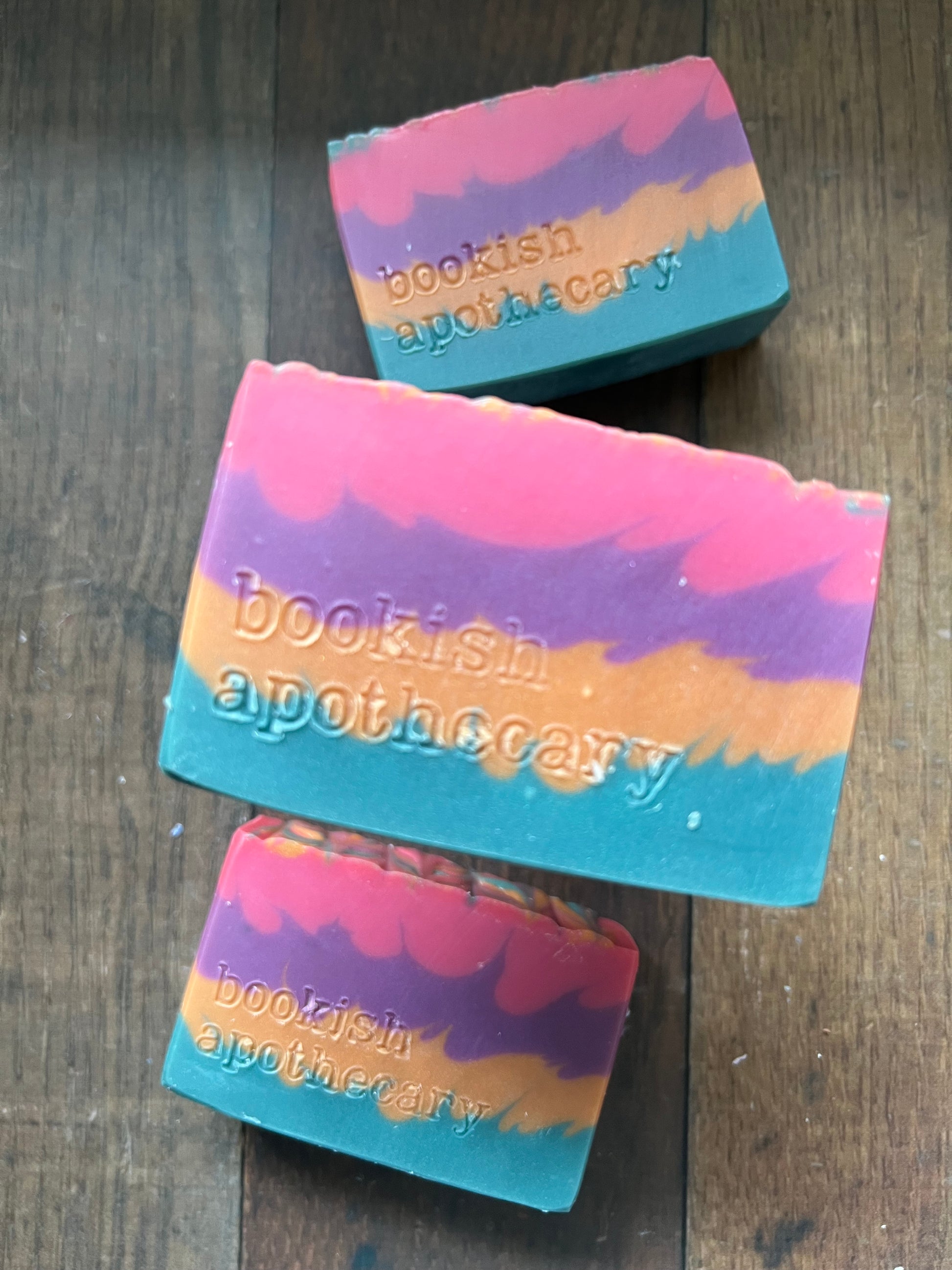 Three colorful soap bars with 'BooKish Apothecary' branding on a wooden surface.