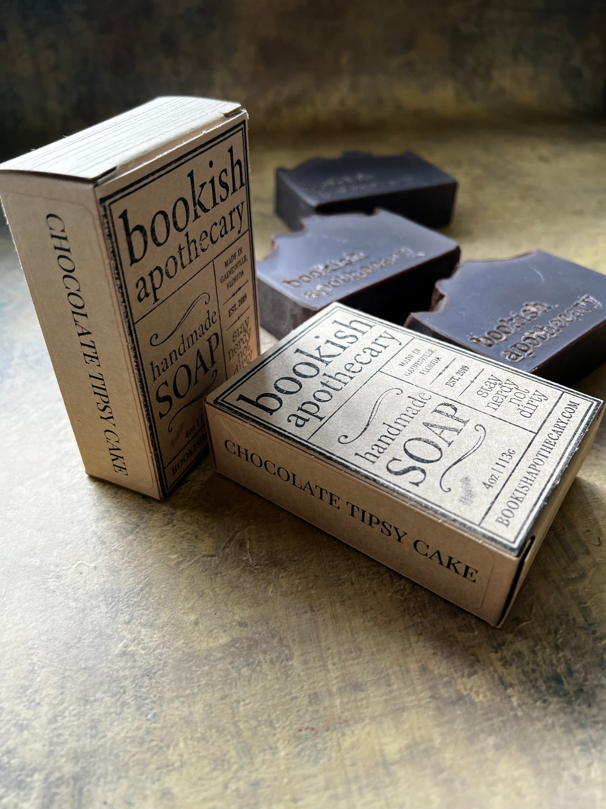 Two vintage-style soap boxes labeled 'bookish apothecary' and 'CHOCOLATE TIPSY CAKE' on a textured metallic surface, with dark soap bars in the background.