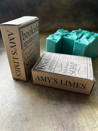 Three teal handmade soap bars with matching kraft paper packaging boxes labeled 'bookish apothecary' and 'AMY'S LIMES' on a rustic metal surface.
