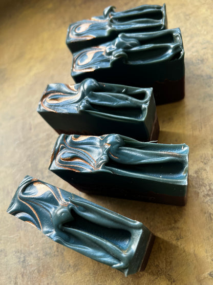 Five rectangular soap bars with a sculpted figure design, featuring a dark teal-green top half with copper-gold marbling and a dark purple bottom half, arranged diagonally on a textured brown surface.