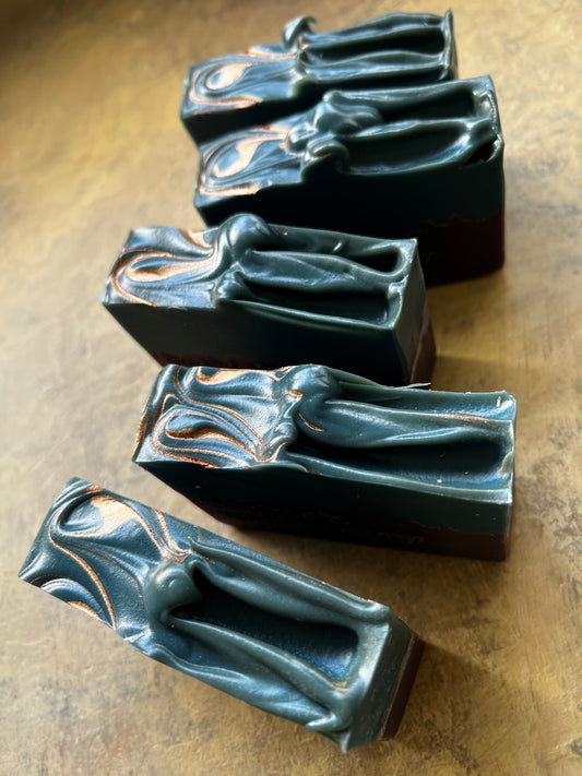 Five rectangular soap bars with a sculpted figure design, featuring a dark teal-green top half with copper-gold marbling and a dark purple bottom half, arranged diagonally on a textured brown surface.