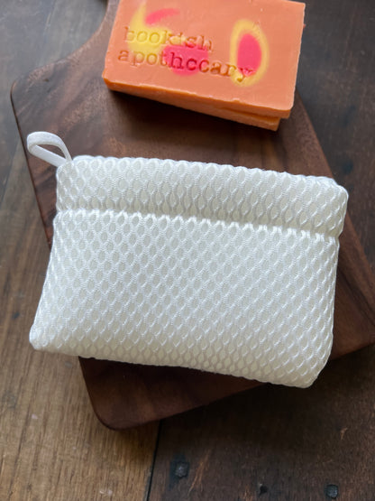 A white soap envelope placed on a wooden surface, with a bar of soap inside it. Other similar soaps envelopes and a colorful bar of soap are visible in the background.