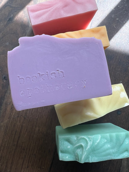 Four bars of soap in different colors on a wooden surface, with 'bookish apothecary' branding.