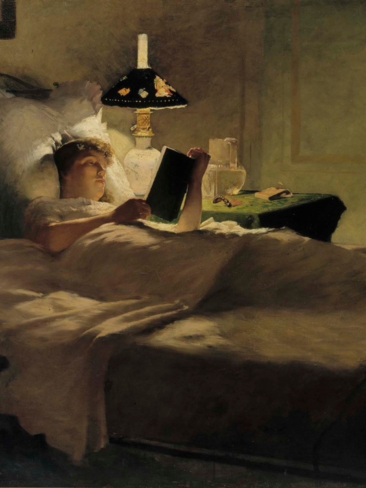 Woman lying in bed reading a book with a lamp and nightstand in the background