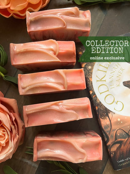 A photo of bars of handmade soap with a pink and white color variation, labeled 'COLLECTOR EDITION' and placed near a book with an ornate cover.