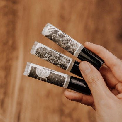 Hand holding three room mist sprays with vintage-style labels against a blurred brown background